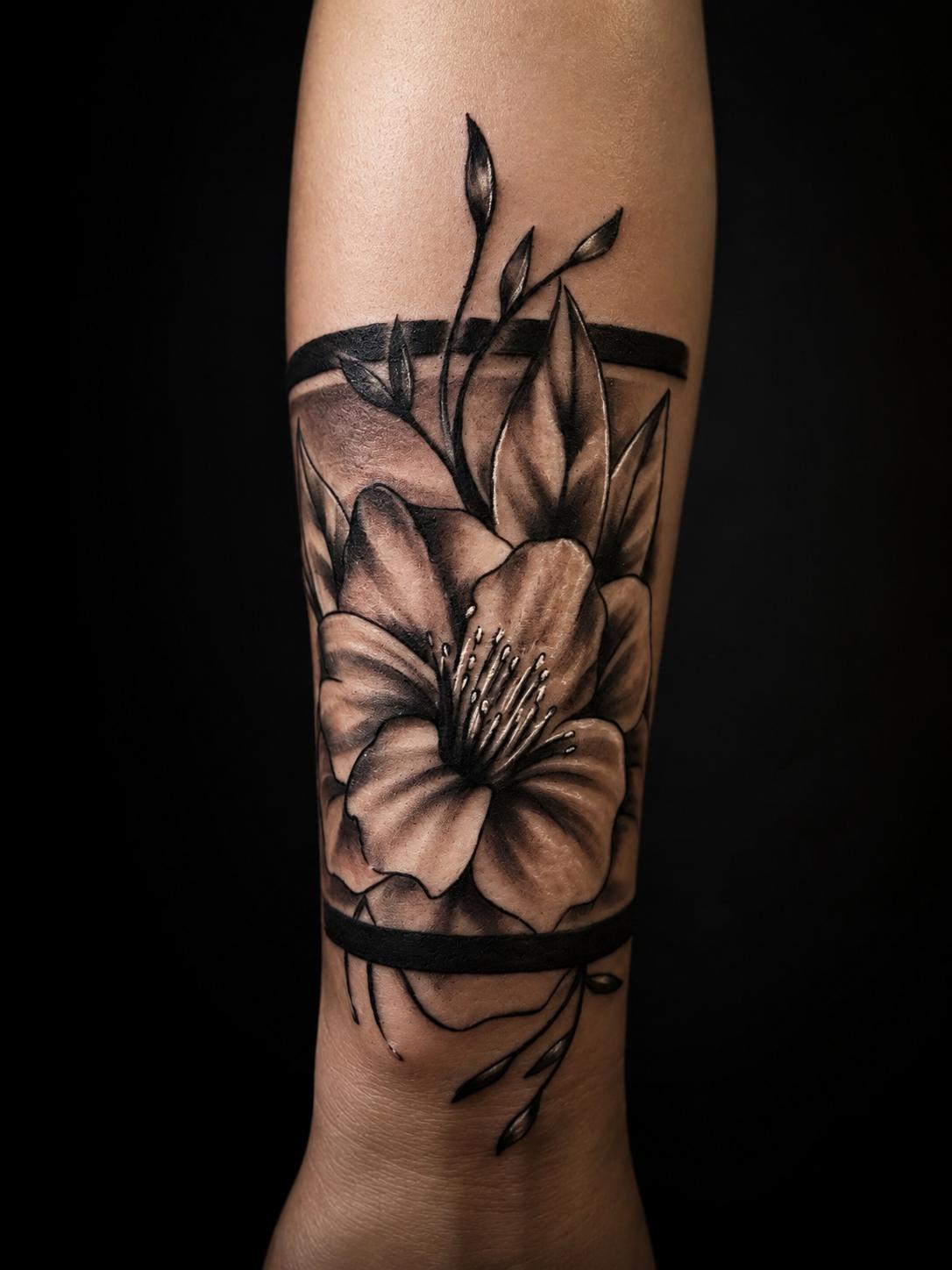 Black-&-Grey-Tattoo einer Blüte zwischen zwei Armbändern am Unterarm